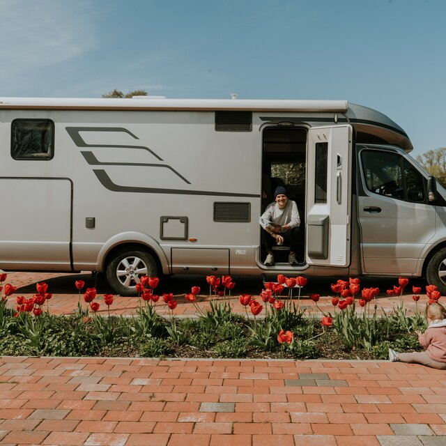 Zijaanzicht van een HYMER-camper met jonge man zittend in open toegangsdeur; rode tulpen op de voorgrond. 