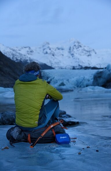 Photo d'un voyage en camping-car en Islande. Un homme en veste verte est assis sur un sac à dos sur une surface glacée, dos au spectateur.