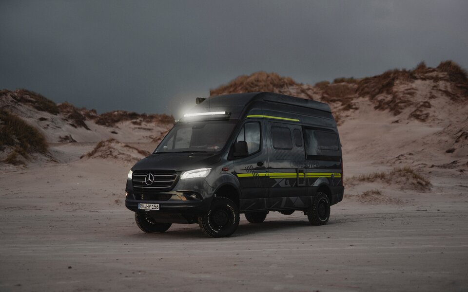 HYMER terränghusbil står i skymningen med strålkastare och ljusramp på en sandstrand med sanddyner i bakgrunden.	