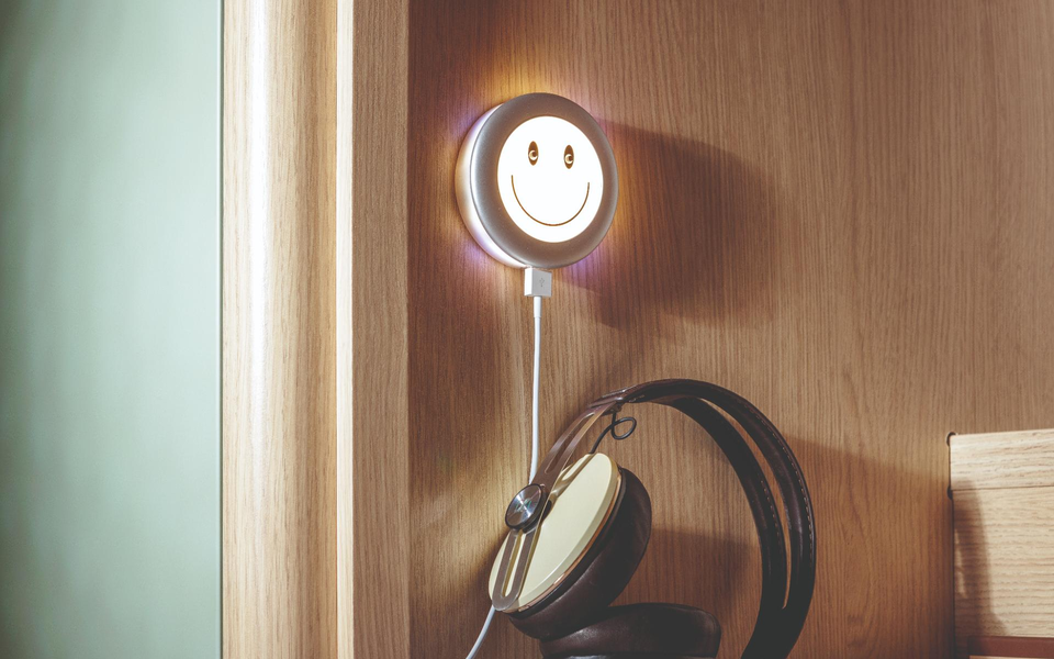 Das Bild zeigt ein leuchtendes Smiley-Wandgerät mit USB-Anschluss, begleitet von Kopfhörern und Smartphone.