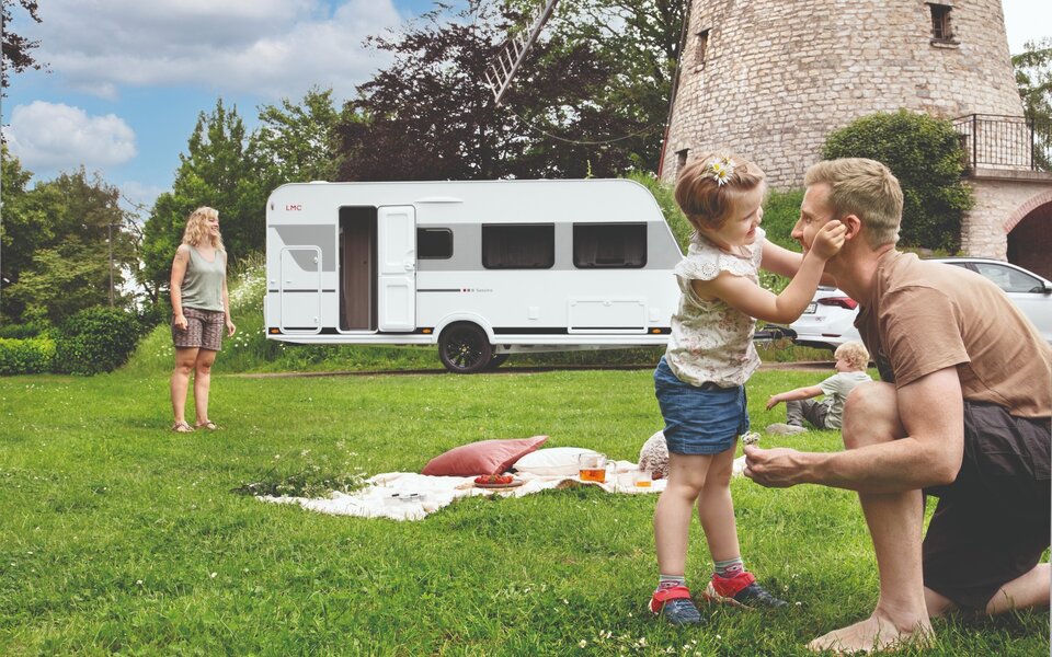 De foto toont een jong gezin dat geniet van een picknick op een groene weide naast de LMC Sassino-caravan en een molen.